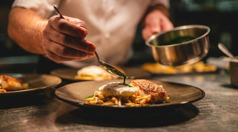Chef plating a dish with fish, pasta, and a poached egg, drizzling green sauce.