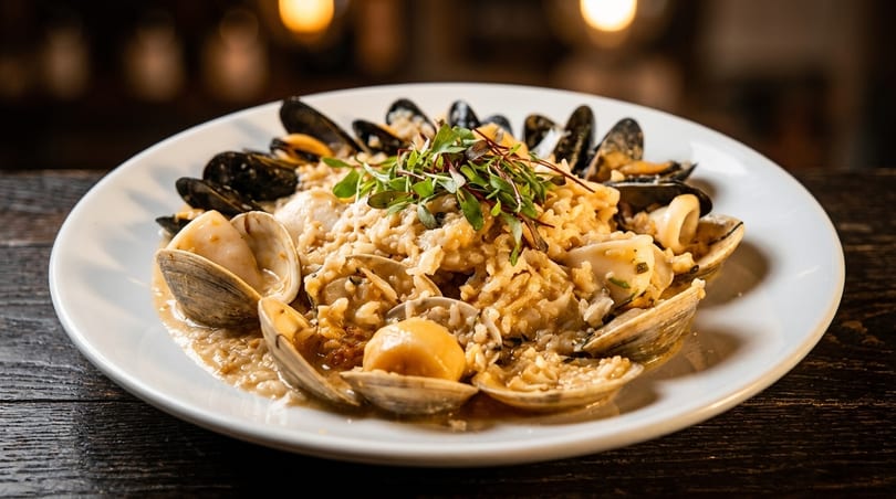 Seafood risotto with mussels, clams, scallops, and squid, garnished with microgreens on a white plate.